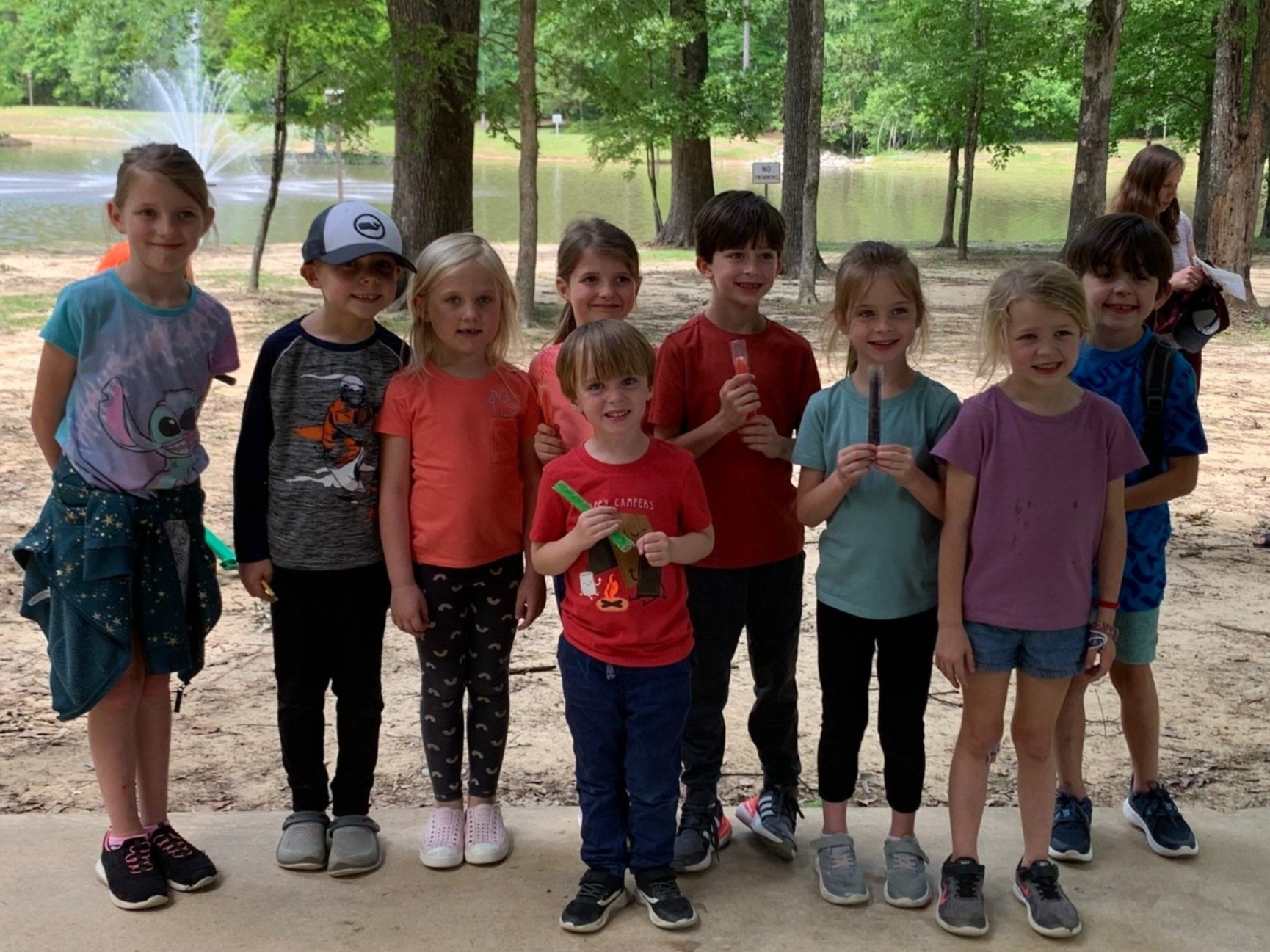 Group of nine children standing in a line outdoors near a pond with a fountain, some holding popsicles.