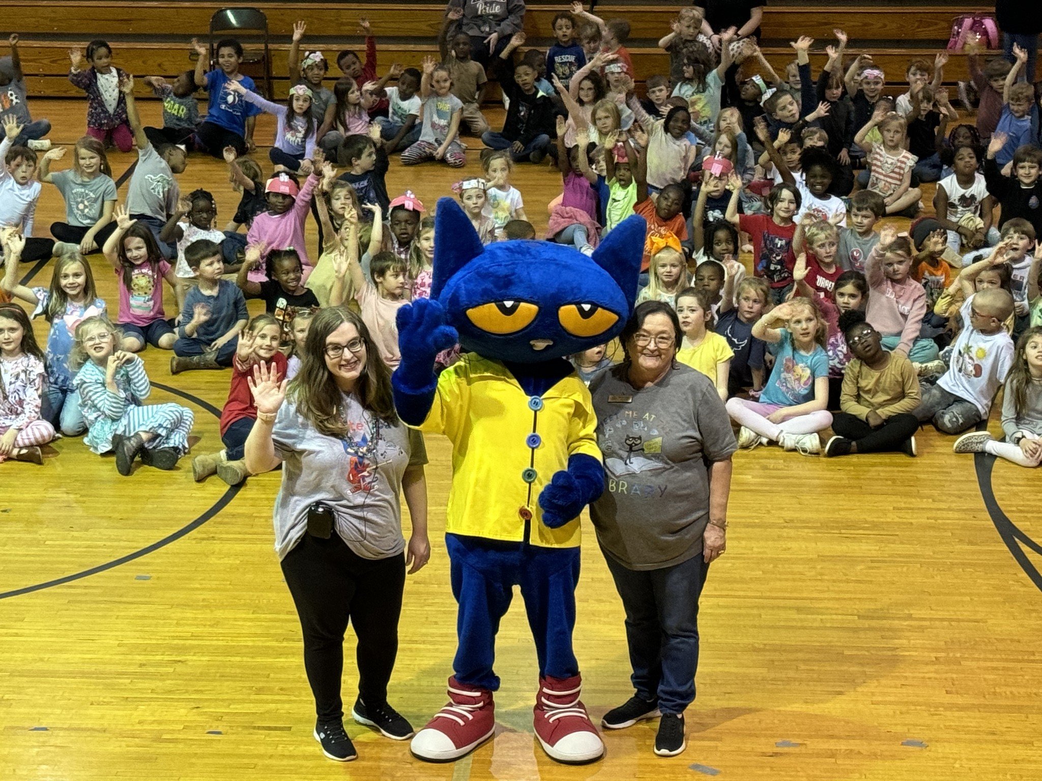 Large group of children in a gymnasium waving at the camera with a blue cat mascot, called Pete, and two adults in front.